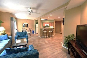 A living room with a sofa, armchair, and a TV on a wooden stand. There is a kitchen area with a microwave and a refrigerator. - Hilton Head Haven, Pool, Free Parking & Beach Access (Hilton Head Island)