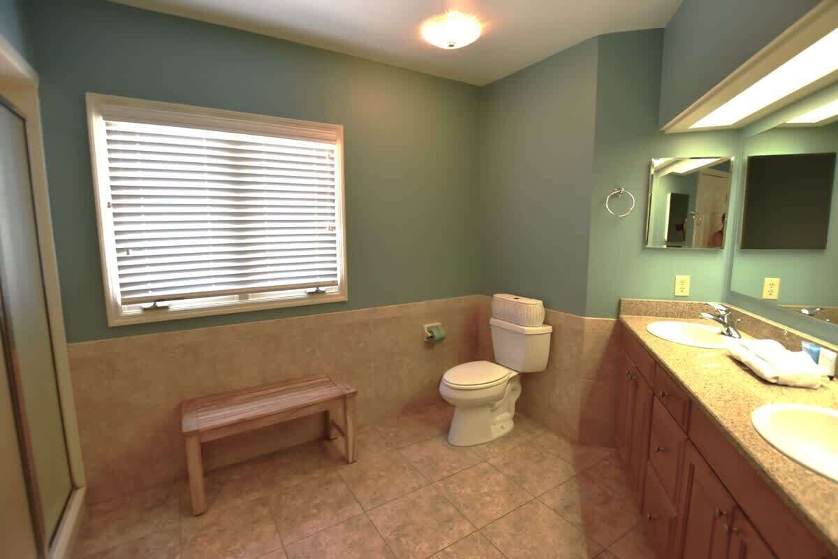Une salle de bain équipée de toilettes, d’un lavabo, d’un miroir et d’une fenêtre avec des stores.