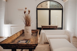 Een kamer met een houten tafel, een vaas met gedroogde planten, een bed met een witte spreidek en een groot boogvenster.