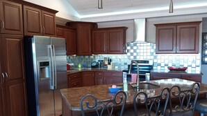 Cocina moderna con armarios de madera, electrodomésticos de acero inoxidable, encimera de granito y una zona de bar con sillas metálicas.