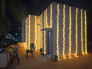 Un edificio con luces colgantes verticales, una puerta y un pequeño espacio de estar con sillas y una mesa.