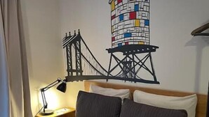 Una camera d'hotel con un letto, un comodino con una lampada, un affresco raffigurante un ponte e una finestra con tende.