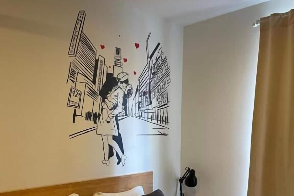 A hotel room with a bed, pillows, a nightstand, a lamp, and a wall mural.