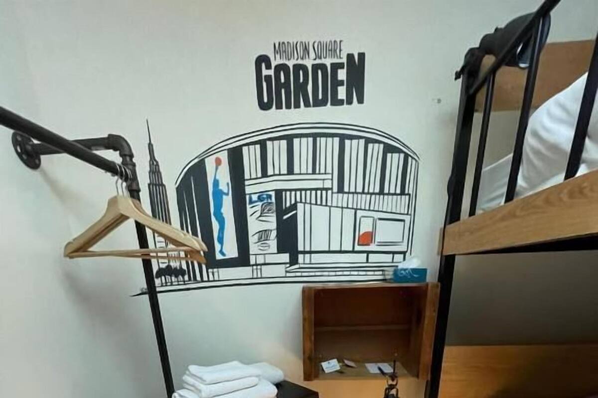 A small, compact room with bunk beds, a compact fridge, a desk with a lamp, and a wall art piece labeled 'Madison Square Garden'.