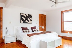 Un dormitorio con cama, mesitas de noche blancas, lámpara de noche, ventilador de techo, ventana y una obra de arte colgada en la pared.