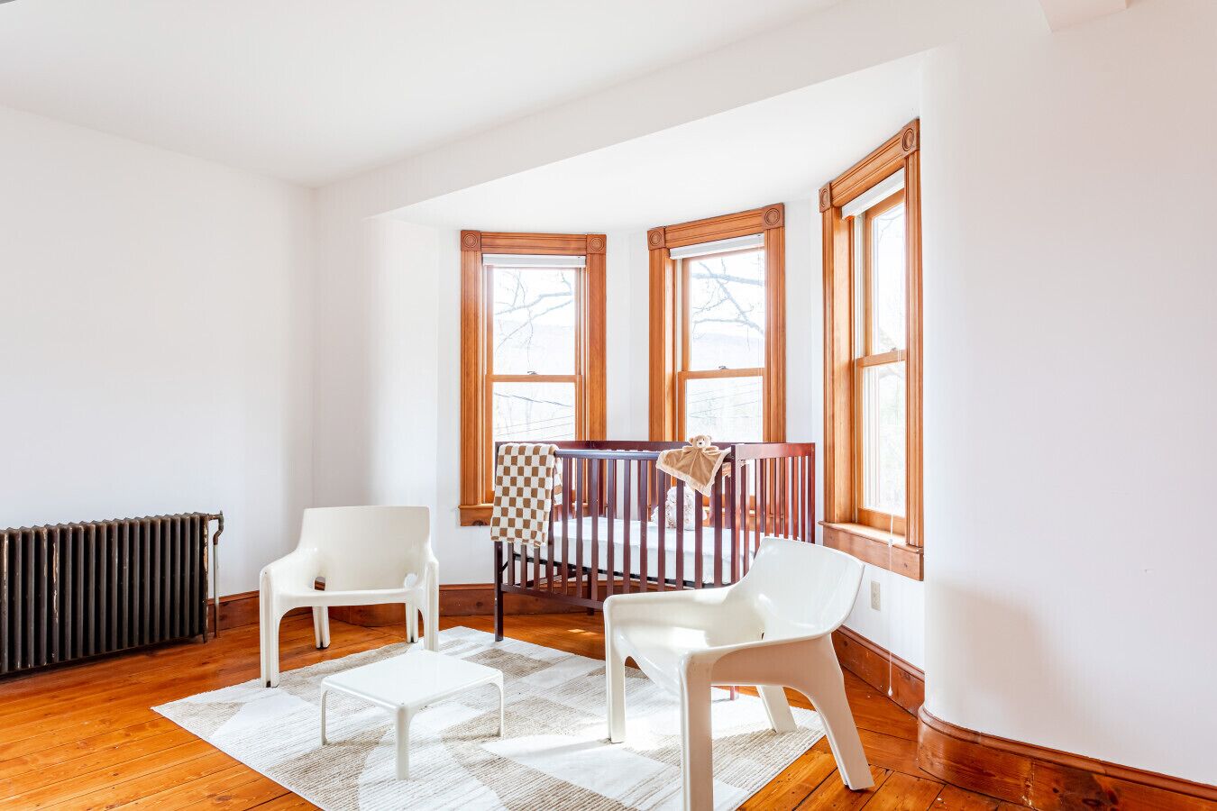 Una habitación con una cuna, dos sillas blancas, un radiador y ventanas amplias.