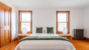 Un dormitorio con cama, dos ventanas, un radiador y suelo de madera.
