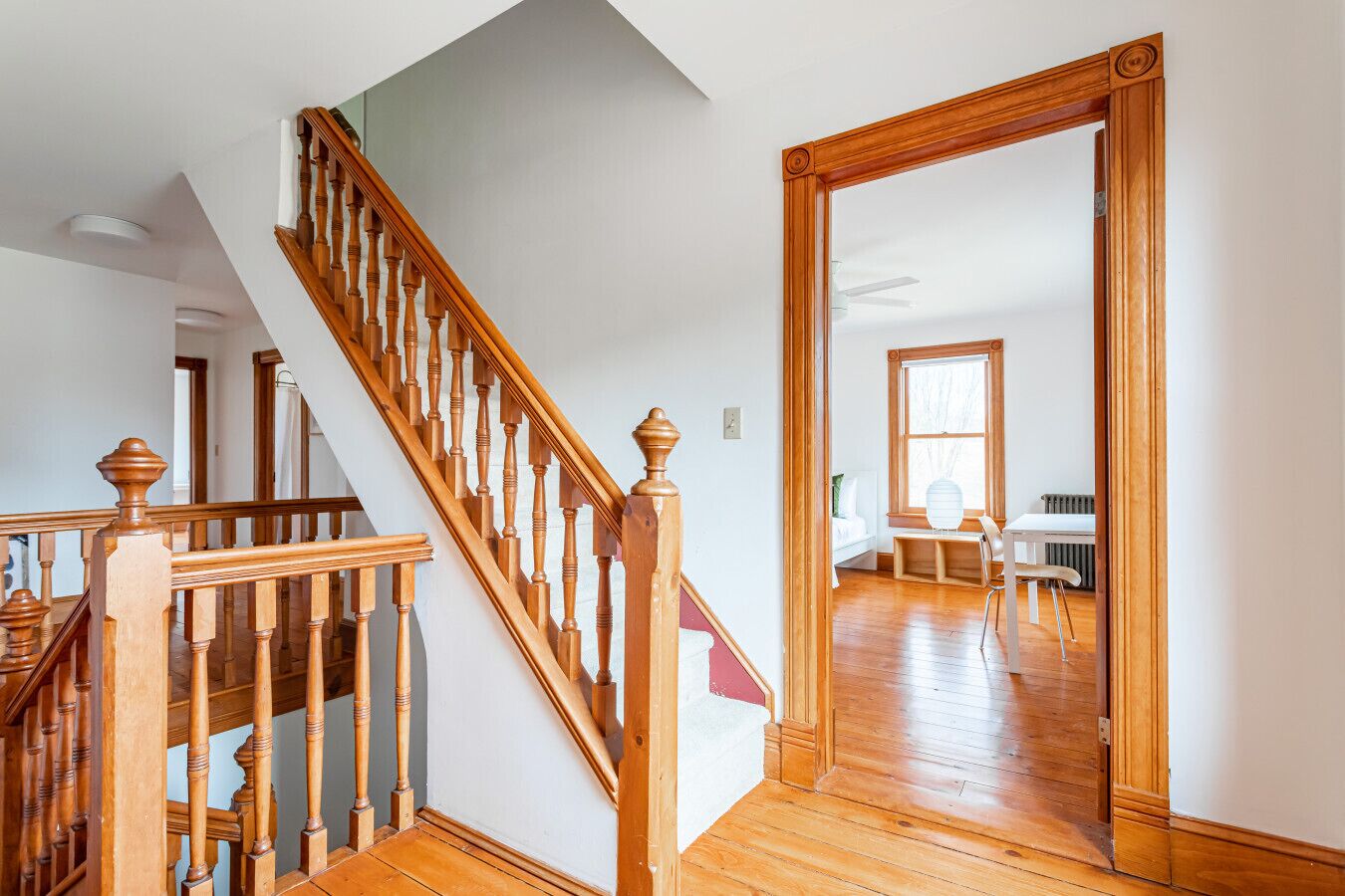 A staircase with wooden balusters leading to a room with a large window, a white radiator, and a white chair.