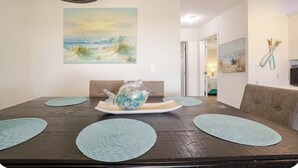 Ein Essbereich mit einem Holztisch, petrolfarbenen Tischsets und einem Gemälde mit Strandmotiv.