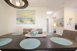 A dining area with a wooden table, teal placemats, and a painting of a beach scene. - Charming 2bd/2ba condo in Gulf Shores/ Walking distance to the beach (Gulf Shores)