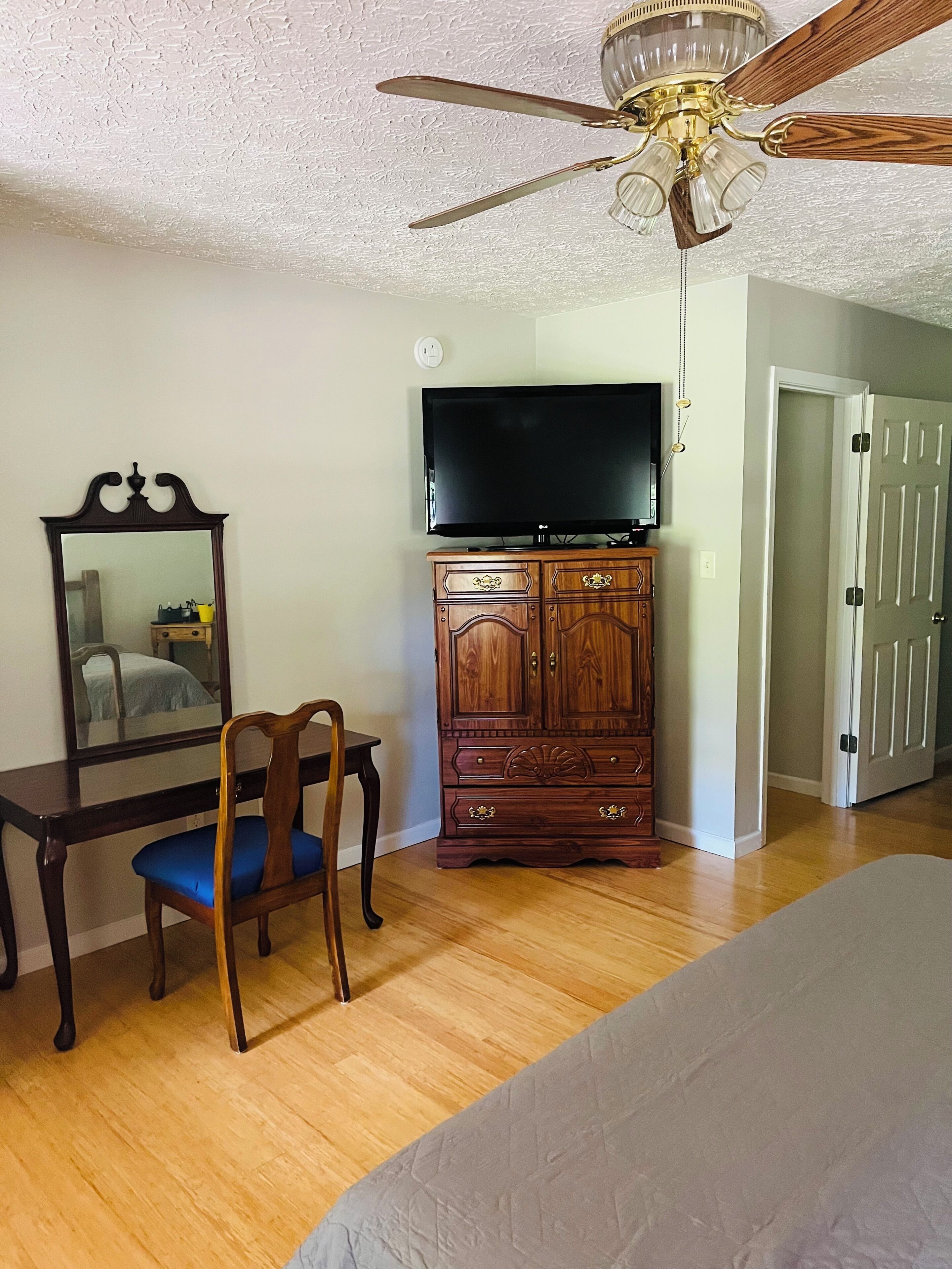 A bedroom with a bed, a wooden dresser, a television, a ceiling fan, and a door.