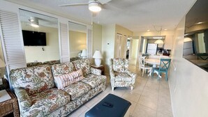 A living room with a sofa, armchair, and ottoman, a dining area with chairs and a table, and a kitchen area with a refrigerator and counter. - Bayfront 2-Bedroom Condo with Private Beach - 314 (Englewood)