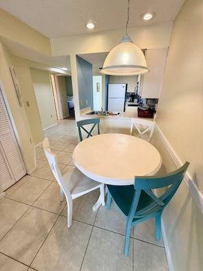 Un comedor con una mesa redonda, cuatro sillas, una lámpara colgante y una cocina al fondo.
