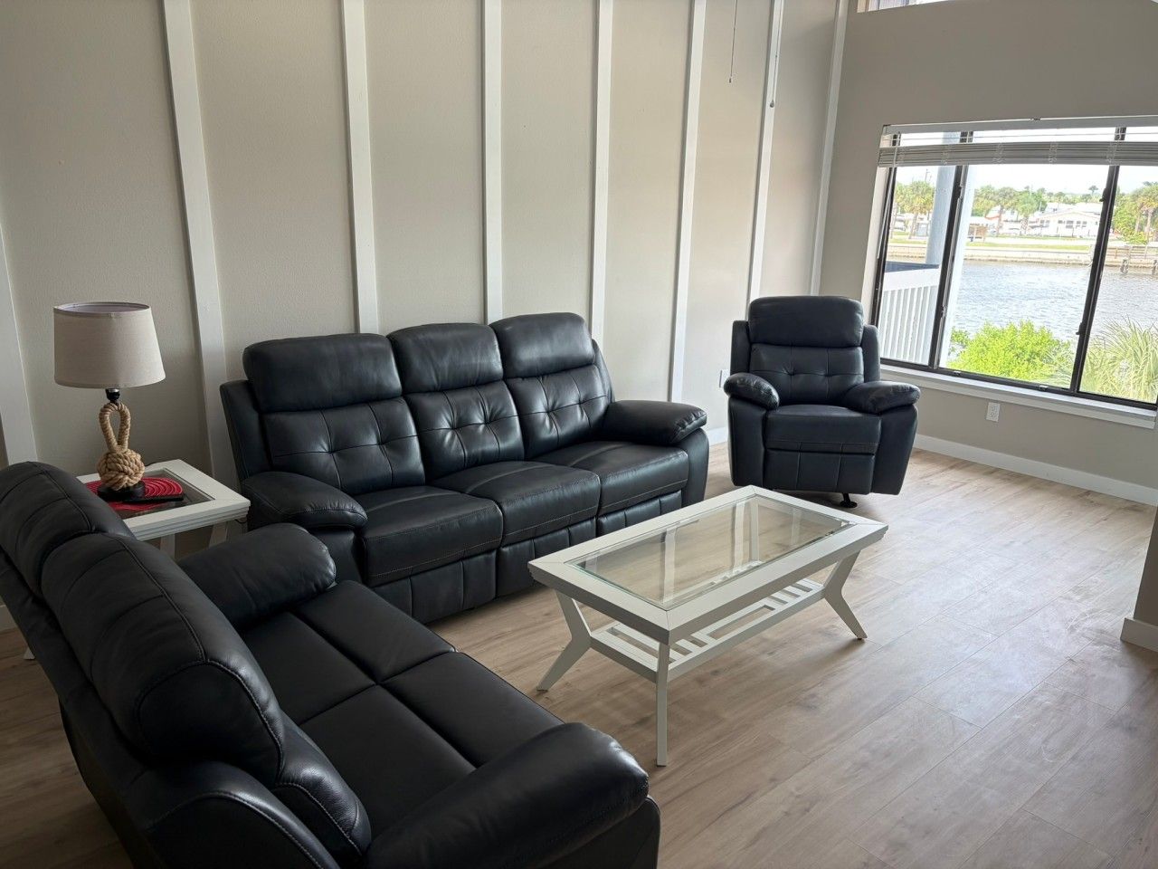 A modern living room with a black leather sofa, a glass coffee table, and a view of a body of water through large windows.