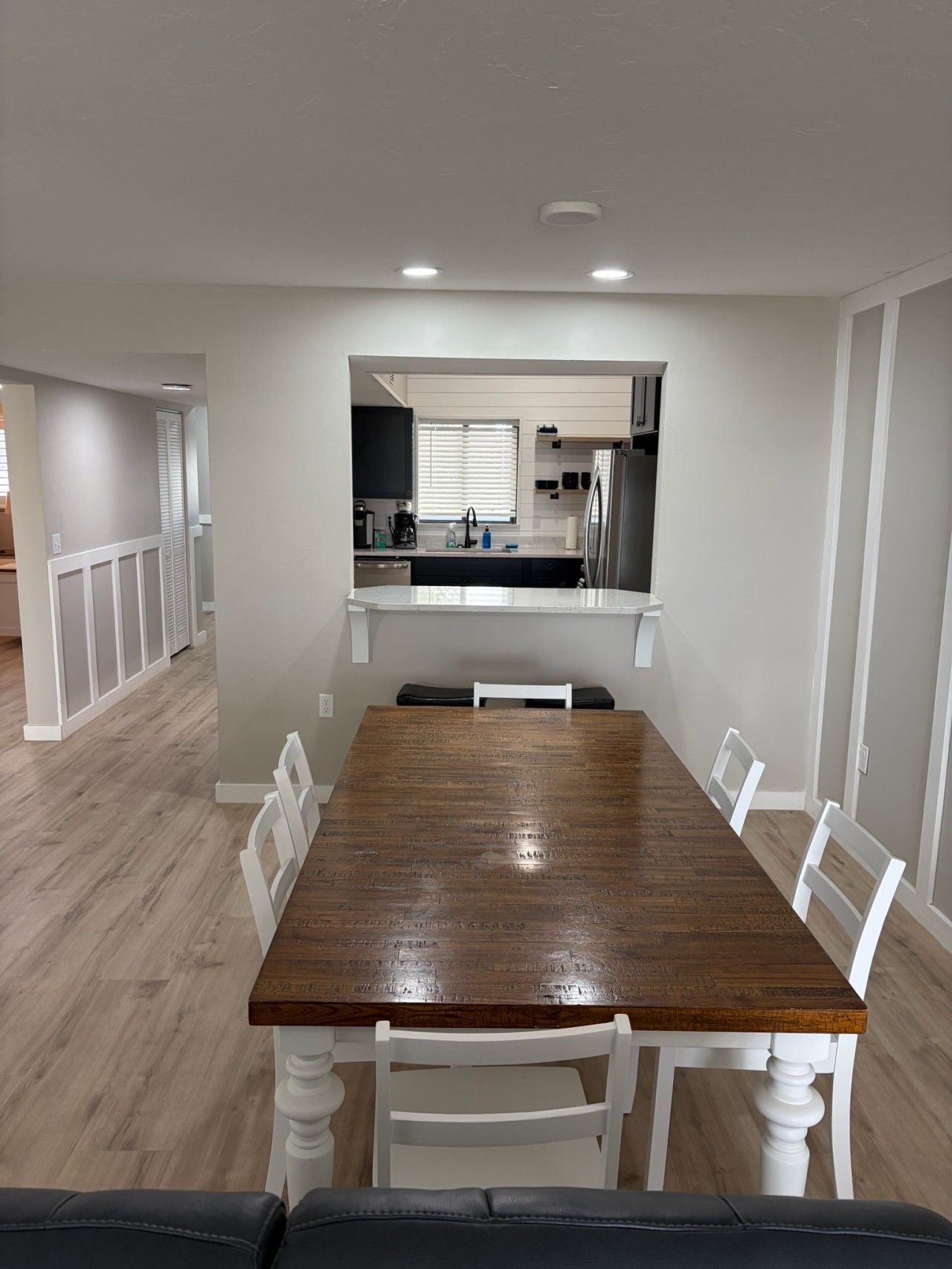 A dining area with a wooden table and white chairs, a kitchen in the background, and a hallway leading to another room.