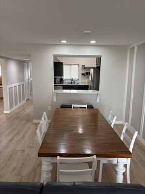 Un coin repas avec une table en bois et des chaises blanches, une cuisine à l’arrière-plan et un couloir menant à une autre pièce.