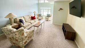 Ein Wohnzimmer mit einer Couch, Sesseln, einem Couchtisch, einer Lampe, einem Fernseher und einem Deckenventilator.
