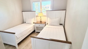 Un dormitorio con dos camas individuales, una ventana con persianas, una mesita de noche con lámpara y suelo de madera.