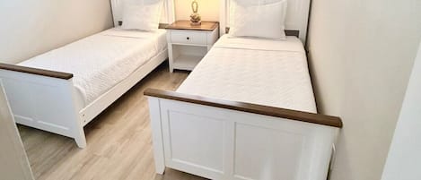 A bedroom with two single beds, a window with blinds, a nightstand with a lamp, and a wooden floor.