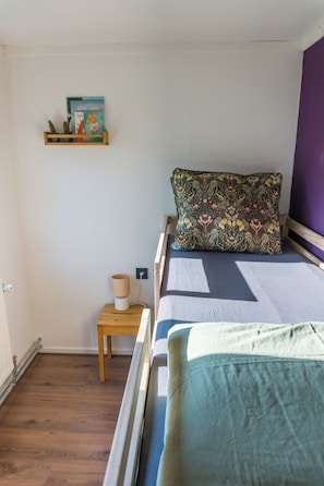 A bedroom with a bed, a bedside table, a lamp, a shelf with books, and a wall-mounted picture.