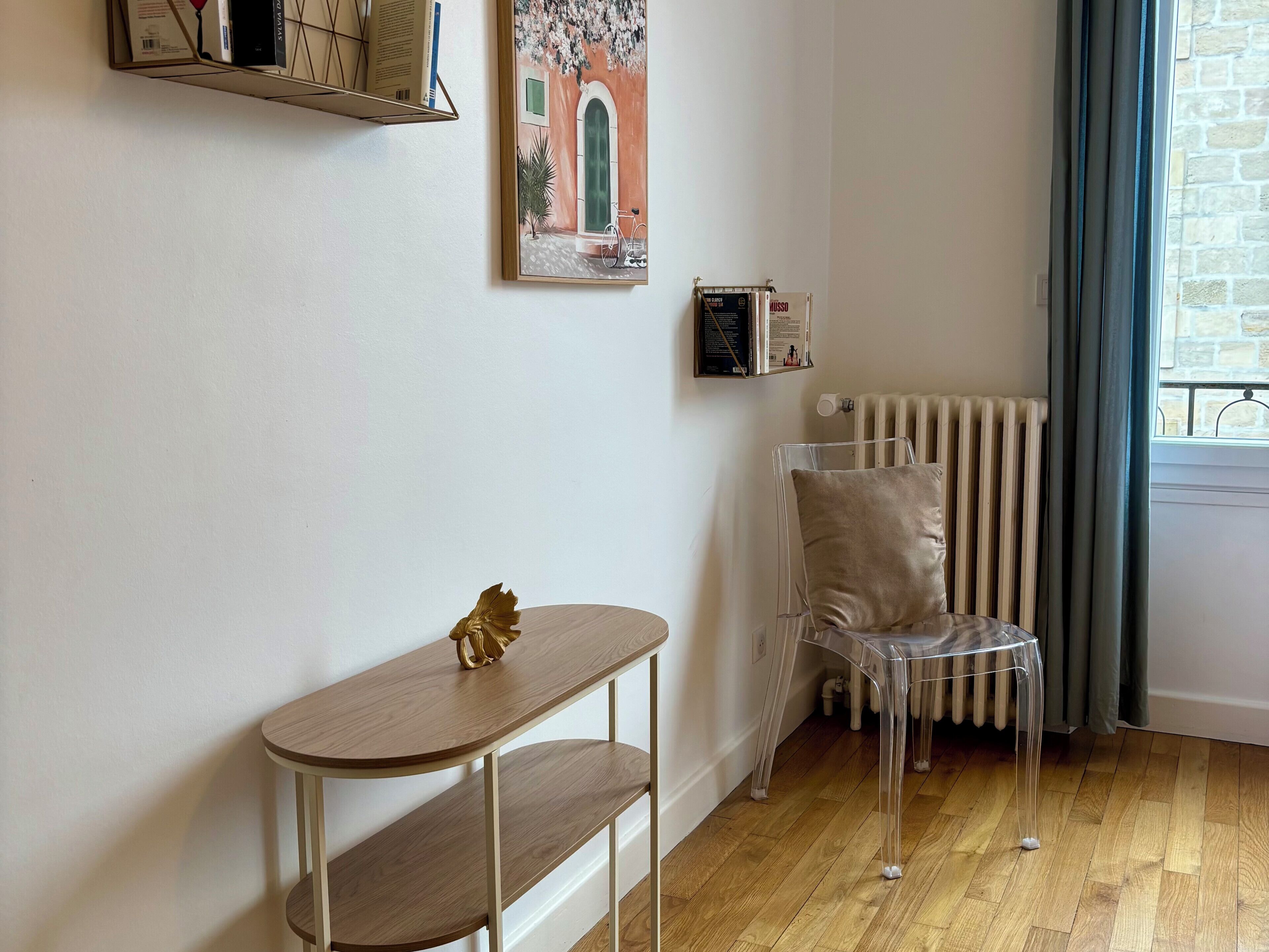 A room with a wooden floor, a clear chair, a small table with a decorative item, a radiator, and a window with curtains.