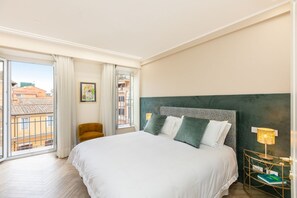 Ein Hotelzimmer mit einem großen Bett, einem Sessel, einem Nachttisch mit Lampe und Blick auf Gebäude durch ein großes Fenster.