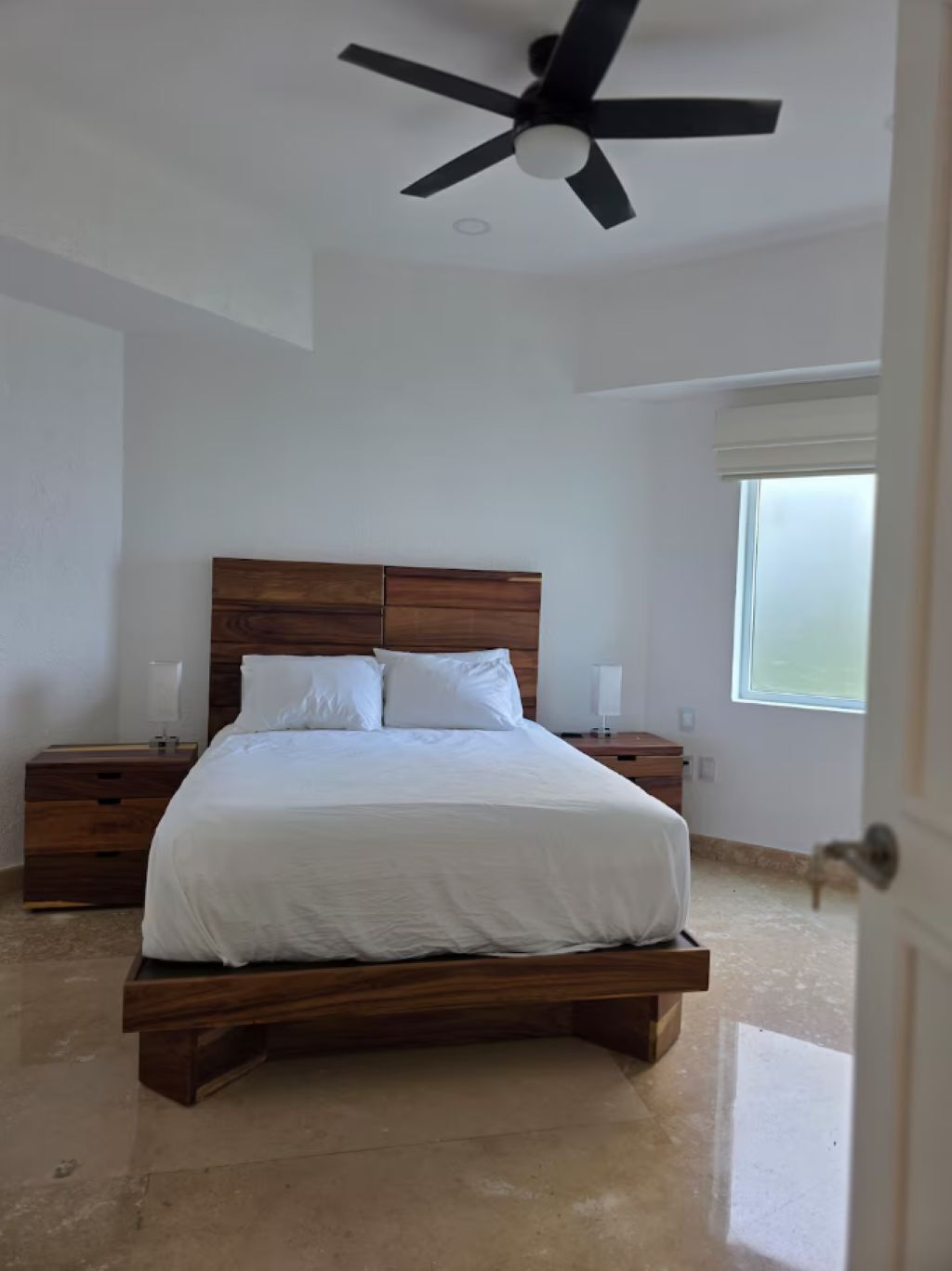 Une chambre avec un lit en bois, du linge de lit blanc, un ventilateur de plafond, des tables de chevet et une fenêtre donnant sur un beau paysage.