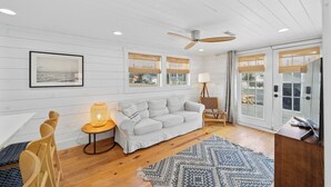 A modern living room with a white wooden floor, a grey sofa, a wooden chair, a lamp, and a ceiling fan.