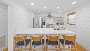 A modern kitchen with white cabinetry, a central island, four wooden chairs, and a window with blinds.