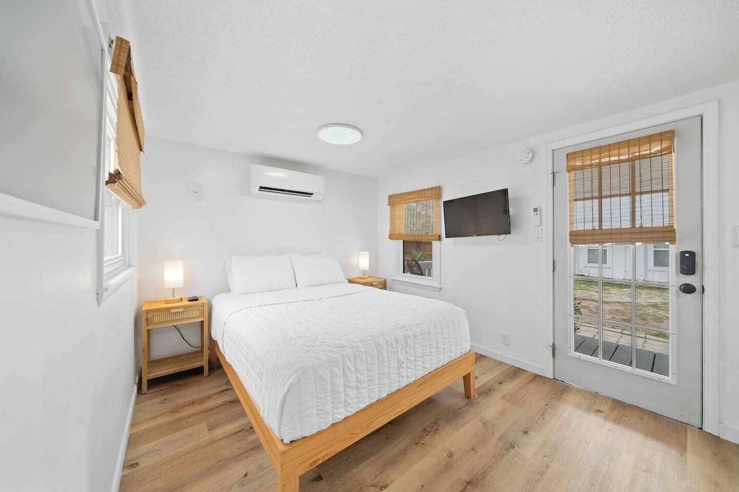 Un dormitorio con una cama de madera, una mesita de noche con lámpara, un televisor montado en la pared y una ventana con persianas.