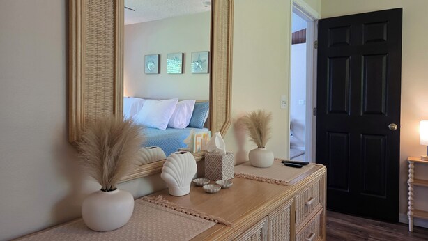 Um quarto com um espelho grande, uma cama com roupa de cama branca, uma cômoda com objetos decorativos e uma porta escura.