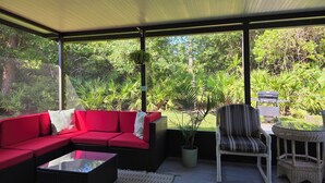 Uma área de estar coberta ao ar livre, com um sofá vermelho, uma cadeira de vime e uma mesa pequena.