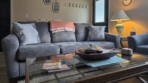Uma sala de estar com um sofá cinza, uma mesa de centro com revistas, uma luminária e quadros nas paredes.