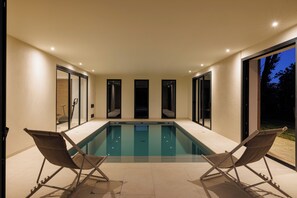Une piscine avec deux chaises longues, un espace clos par des parois de verre et une vue sur l’extérieur.