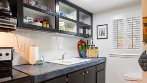 Una cocina con encimera negra, salpicadero blanco de azulejos, un fregadero y una ventana con persianas.