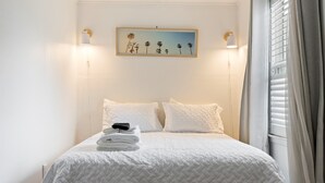 Una cama bien hecha con ropa blanca, un cuadro de palmeras en la pared y una ventana con persianas.