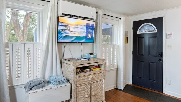 Una habitación con una televisión montada en la pared, una mesita de mimbre con libros, una puerta y una ventana con contraventanas.