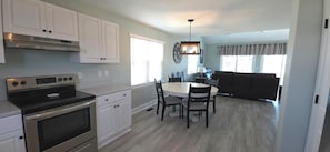 A compact kitchen with white cabinets, a stove, and a dining area with a table and chairs. - 2nd Row, Ocean views. 3br. (sleeps 8) -large deck, Surf City NC, Steps to beach  (Surf City)