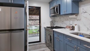 Una cucina con mobili blu, elettrodomestici in acciaio inossidabile, una schiena di marmo e vista su uno spazio esterno attraverso una porta in vetro.