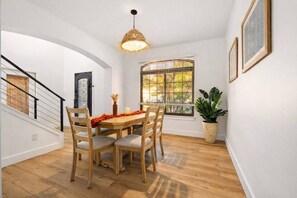 Un coin repas avec une table en bois, des chaises, un luminaire suspendu, une fenêtre avec des stores et une plante en pot.