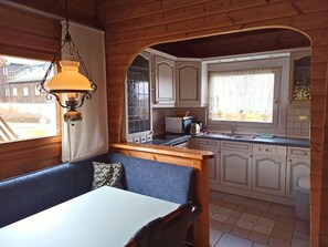 Een gezellig chaletinterieur met een eethoek, houten wanden, een keuken met witte kastjes en een raam met uitzicht op een tuin.