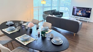 A modern living room with a dining table set for four, a grey sofa, and a television displaying a program.