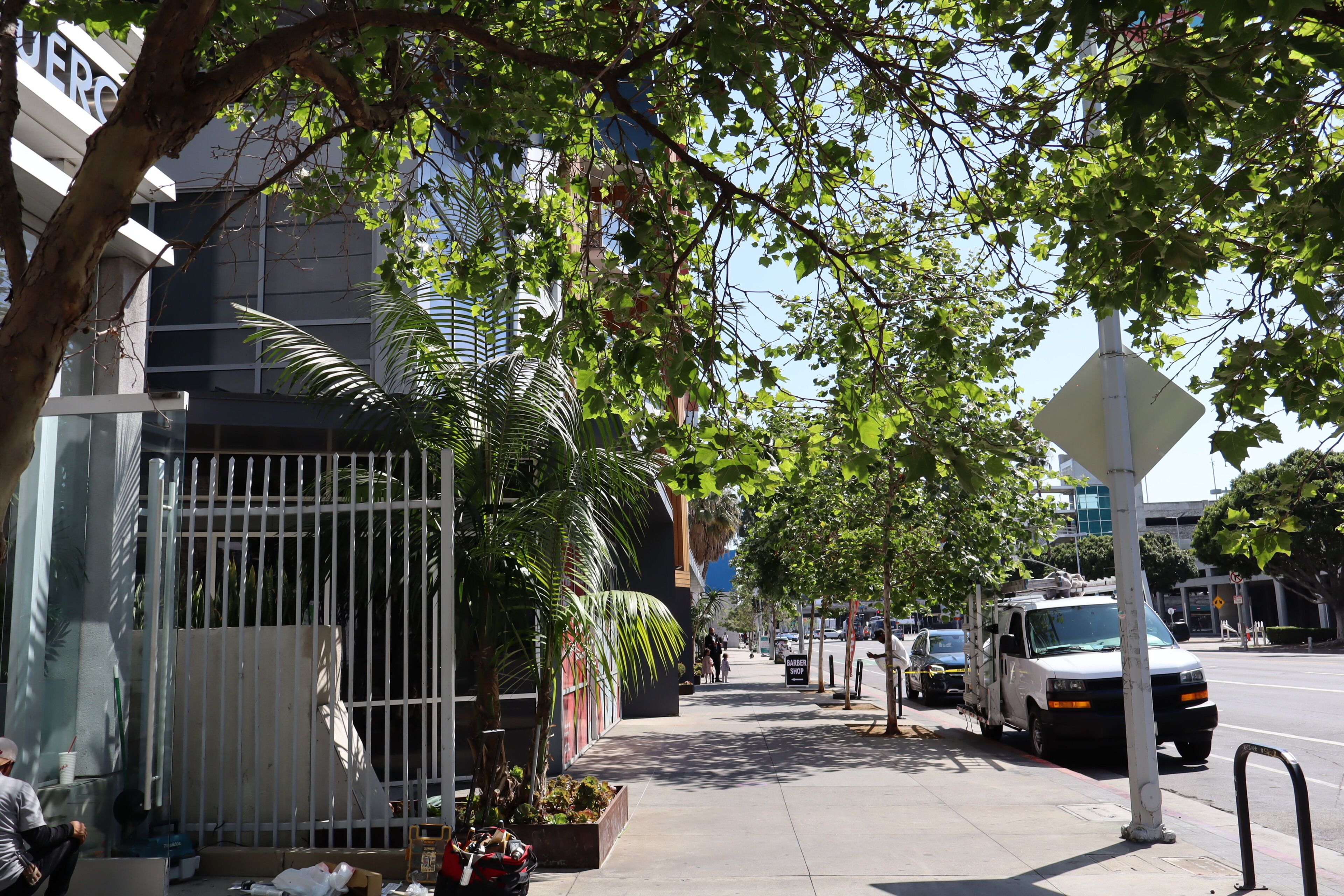 A sidewalk lined with trees and parked vehicles, a gated entrance, and a building with a sign.