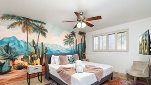 A bedroom with a bed, a ceiling fan, a nightstand, a chair, a window with blinds, and a mural on the wall. - Paradise Palms - Scottsdale 4BR w/ Heated Pool, Fire Pit, PingPong (Scottsdale)