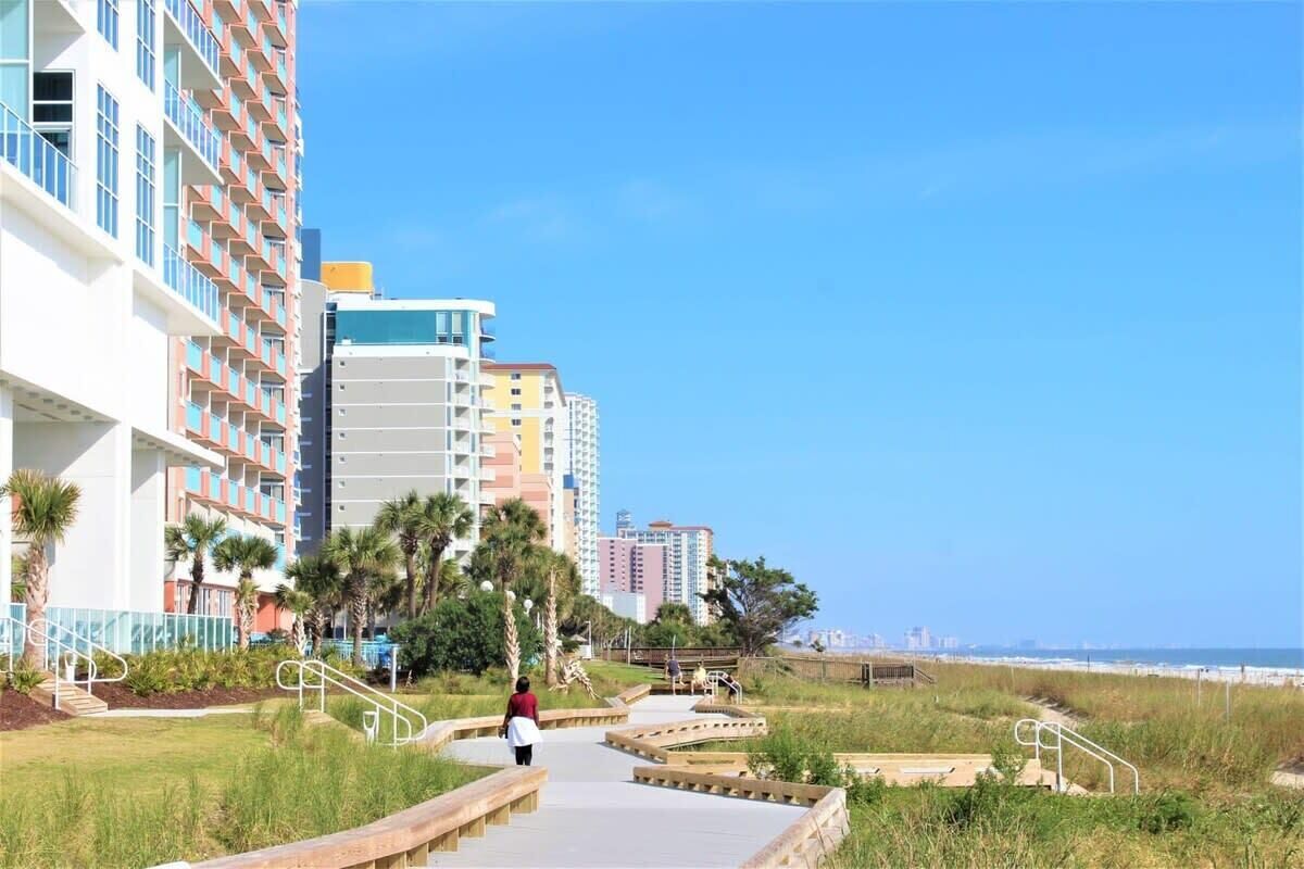 A coastal walkway with a person walking, surrounded by buildings and palm trees.