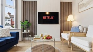 A living room with a TV displaying 'NETFLIX,' a blue sofa, a white sofa, a round coffee table with fruits, and a lamp. - Modern 3BR | Parking | Work trips | Relocators | Events | Flexible stays (Manchester)