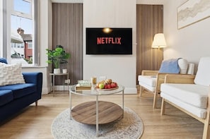A living room with a TV displaying 'NETFLIX,' a blue sofa, a white sofa, a round coffee table with fruits, and a lamp. - Modern 3BR | Parking | Work trips | Relocators | Events | Flexible stays (Manchester)
