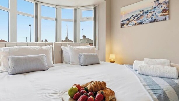 A hotel room with a large bed, a view of a coastal town, and a breakfast tray with fruits and pastries. - Modern 3BR | Parking | Work trips | Relocators | Events | Flexible stays (Manchester)