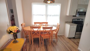 Zona pranzo con tavolo e sedie, una lampada, una cucina completa di elettrodomestici e una finestra con tende.