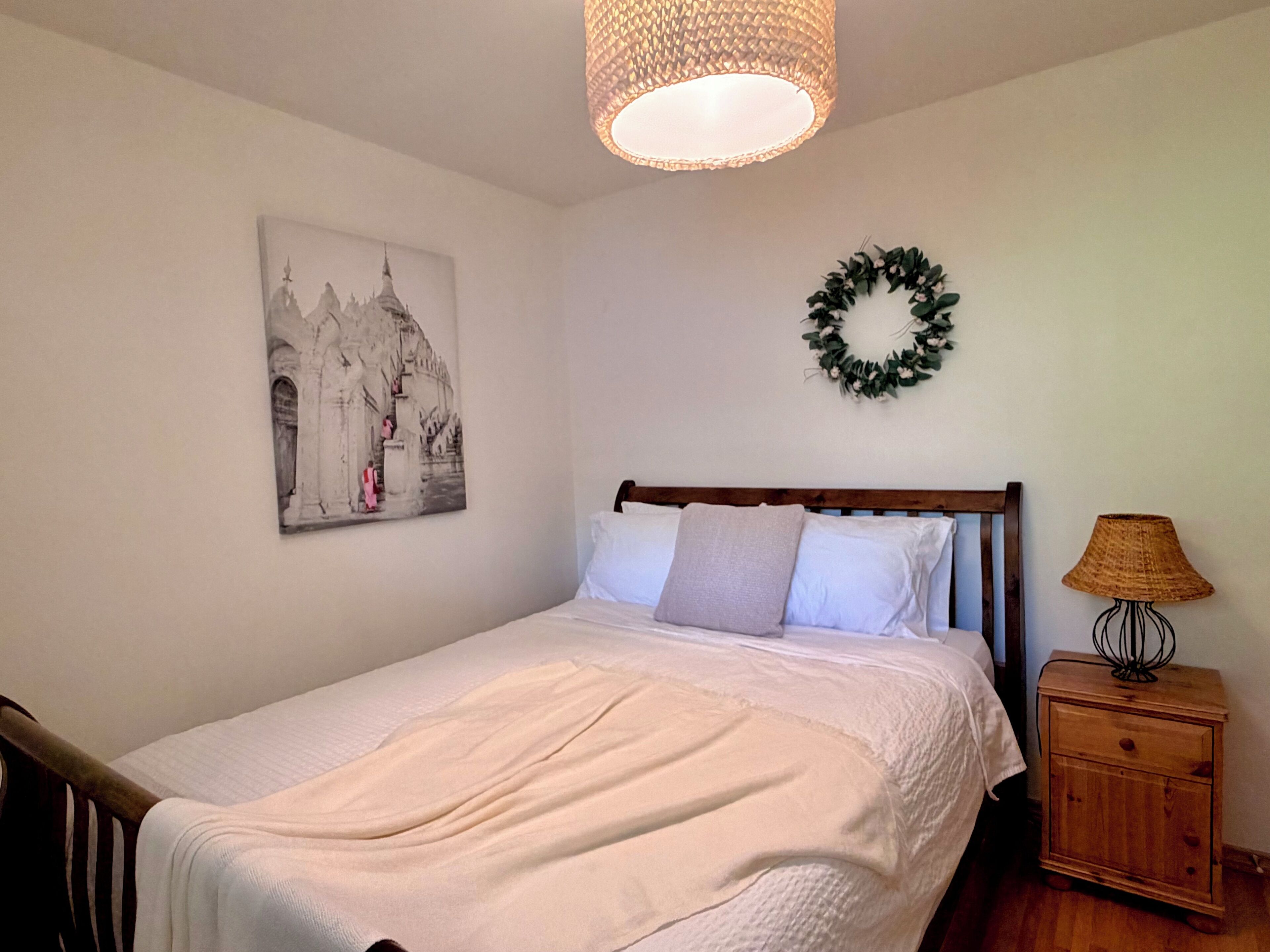 Een slaapkamer met een bed, nachtkastje, lamp, wandlamp, schilderij en krans.